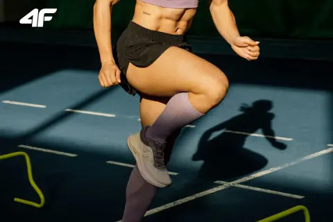 Kobieta wykonuje ćwiczenia na kolano biegacza na bieżni. Widoczne są jej nogi w skarpetach kompresyjnych i buty sportowe.