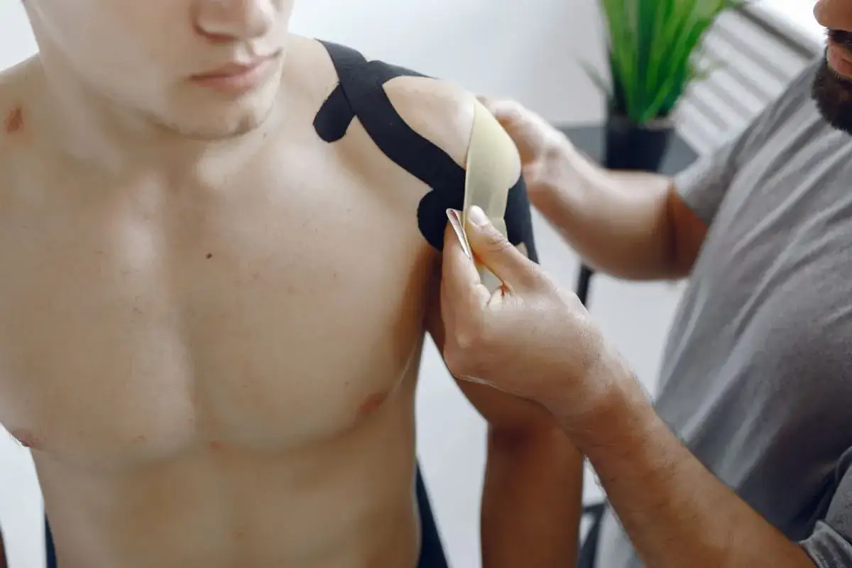 Fizjoterapeuta zakłada taśmy kinezjotaping na bark pacjenta. Długo utrzymuje się ból po artroskopii barku? Taśmy mogą pomóc.